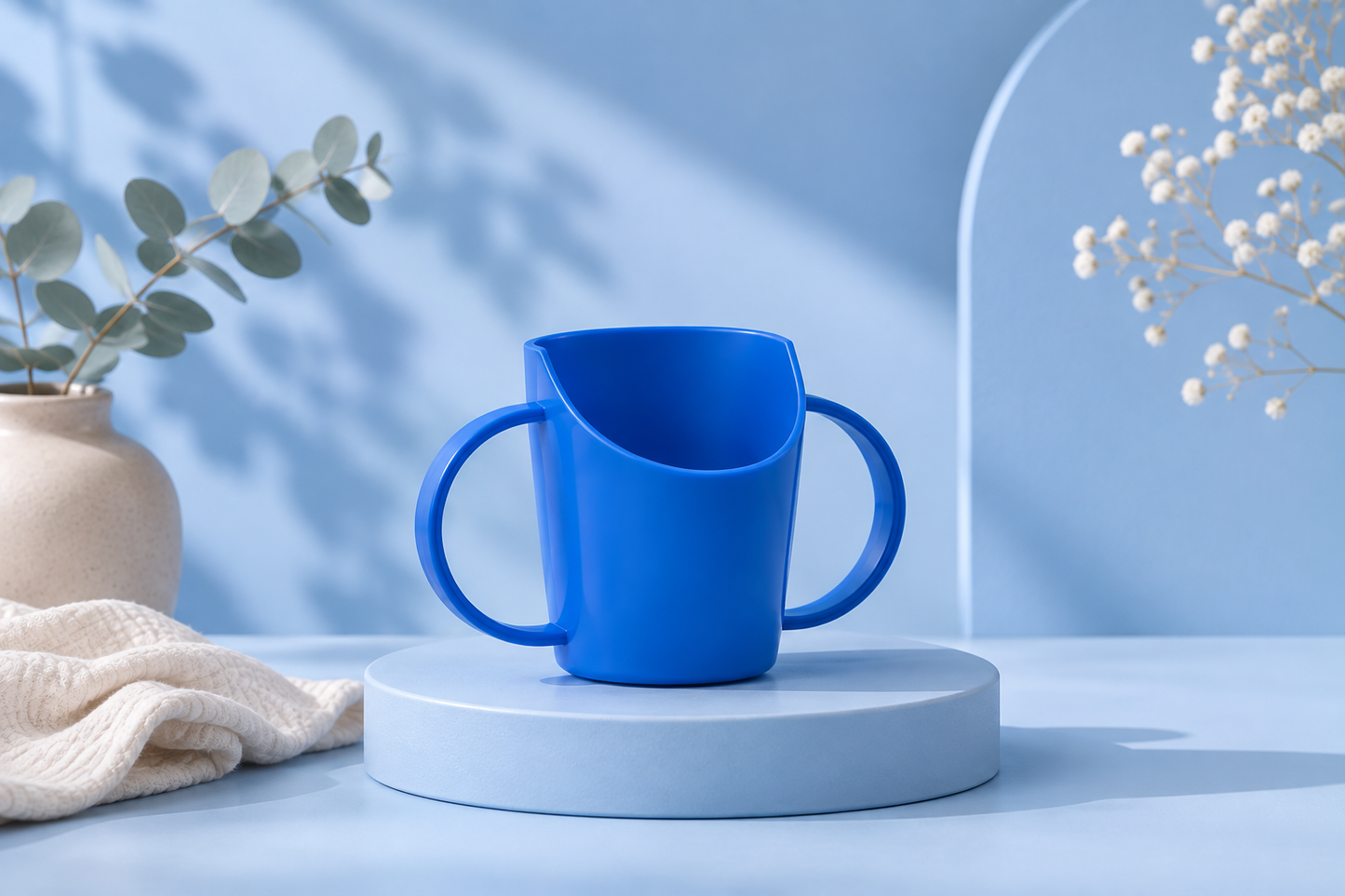 A bright blue kids friendly mug with a unique asymmetrical slanted rim and double handles, displayed on a round pedestal against a light blue background with decorative plants.