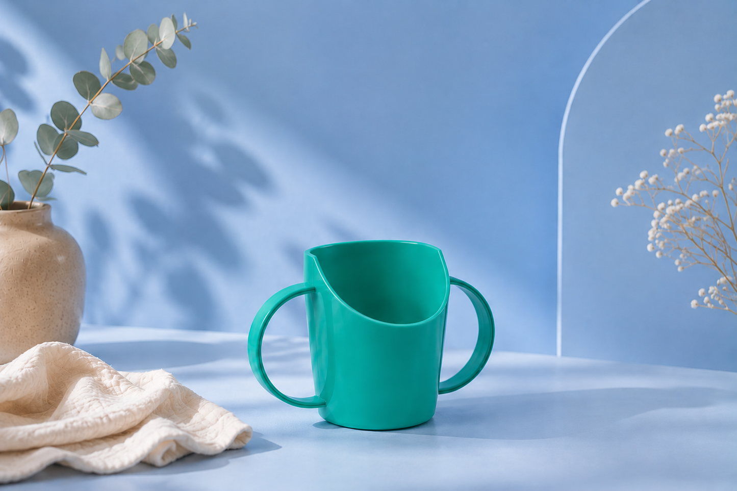 A teal child's cup with a wide mouth and two loop handles, styled on a light blue surface with a beige cloth, a vase with eucalyptus, and dried flowers against a blue background.
