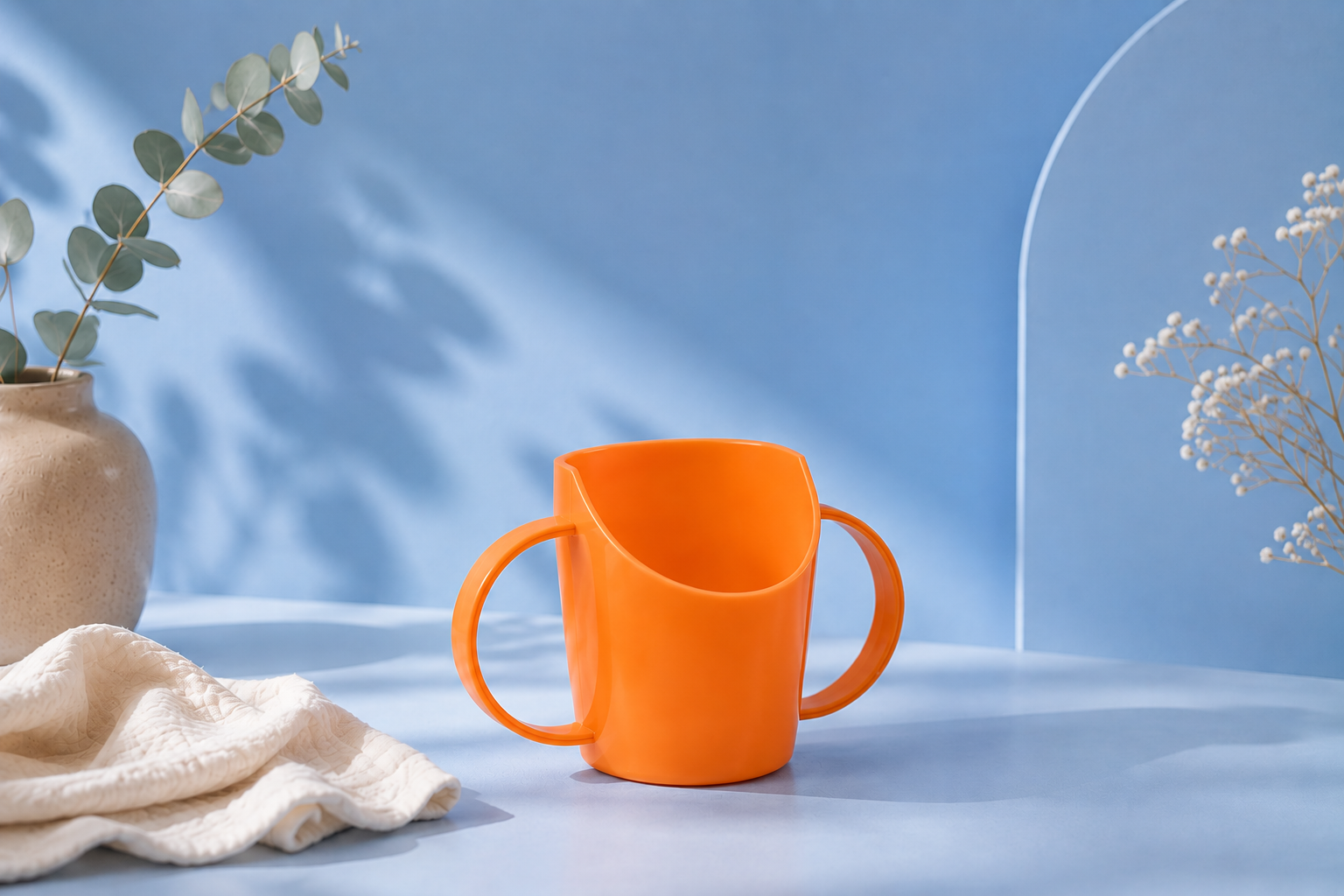 An orange child's cup with two handles, styled on a light blue surface with a beige cloth, a vase with eucalyptus, and dried flowers against a blue background.